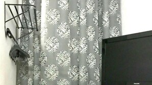 Una habitación con una televisión sobre un aparador de madera, una ventana con cortinas estampadas y un zapatero en la pared.