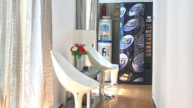 A hallway with a white chair, a small table with a vase of flowers, and a vending machine.