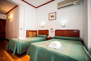 Habitación de hotel con dos camas individuales, cada una con cubrecama verde y una toalla blanca sobre la cama. Ambas camas tienen cabecero de madera, y una mesita de noche con lámpara. El suelo es de madera y la pared blanca tiene un cuadro enmarcado encima de las camas.