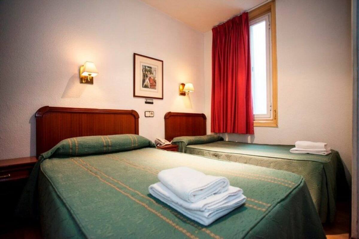 A hotel room with two single beds, each with a green bedspread and a white towel on the bed. There are two wall-mounted lamps and a framed picture on the wall.