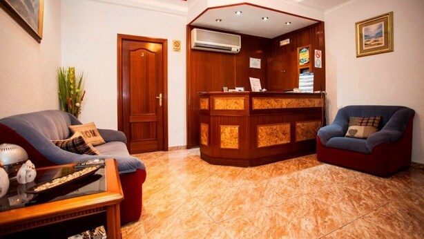 A reception area with a wooden reception desk, a blue sofa, a red sofa, and a glass coffee table. - Pensión Carrera (Madrid)