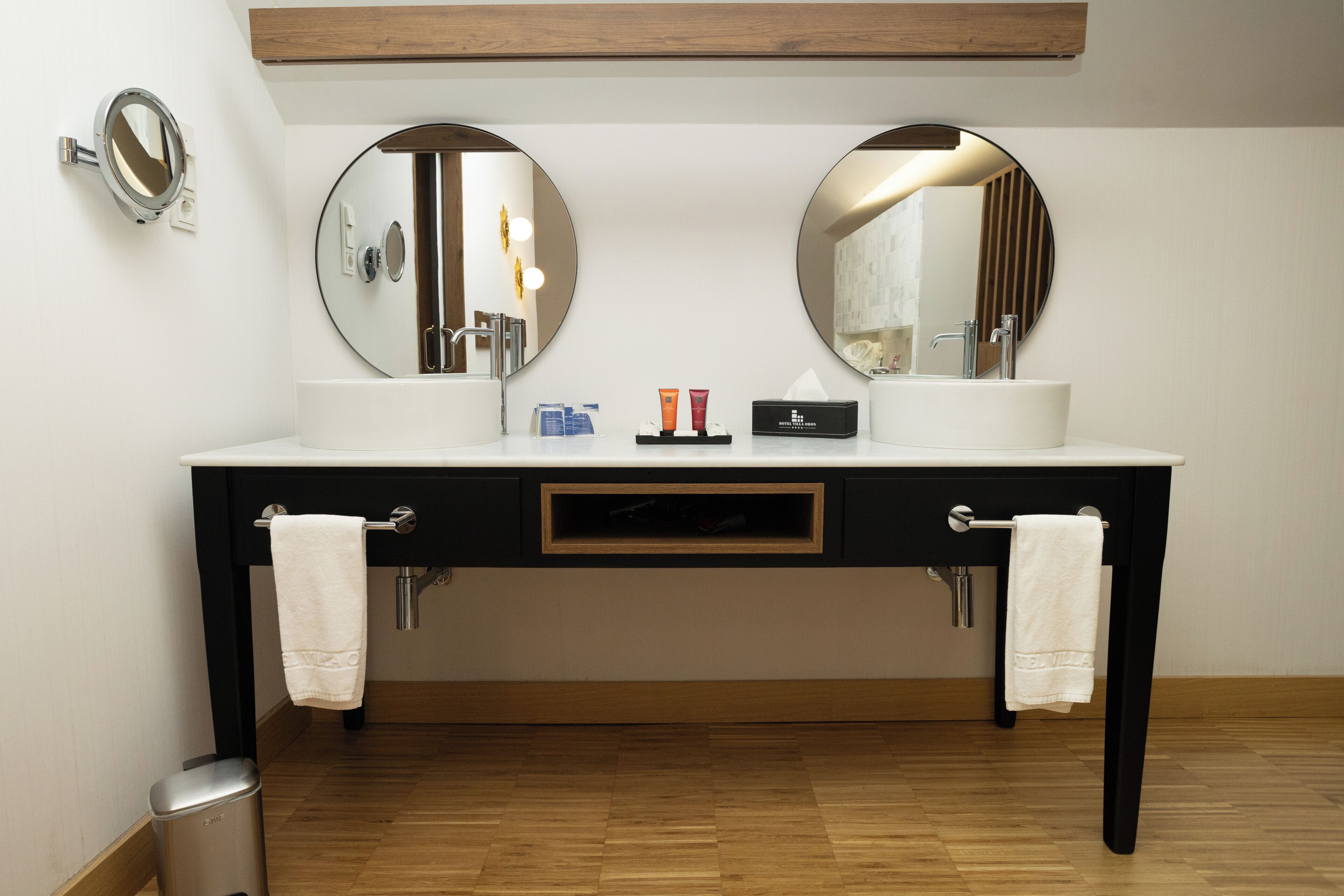 Un baño con tocador negro, dos espejos redondos, lavamanos blanco y suelo de madera.