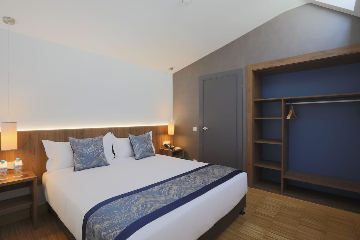 A modern hotel room with a large bed, bedside table with a lamp, a wardrobe, and a nightstand.