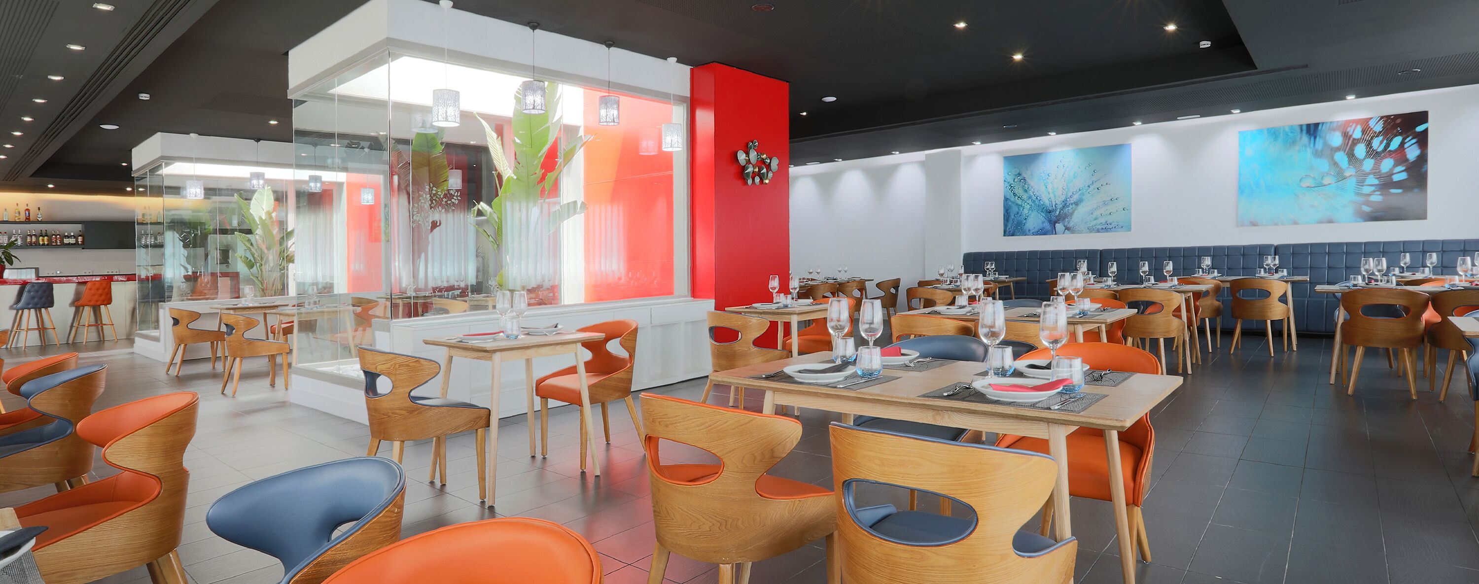 A modern restaurant with wooden tables and orange chairs, a red accent wall, and abstract art on the walls.