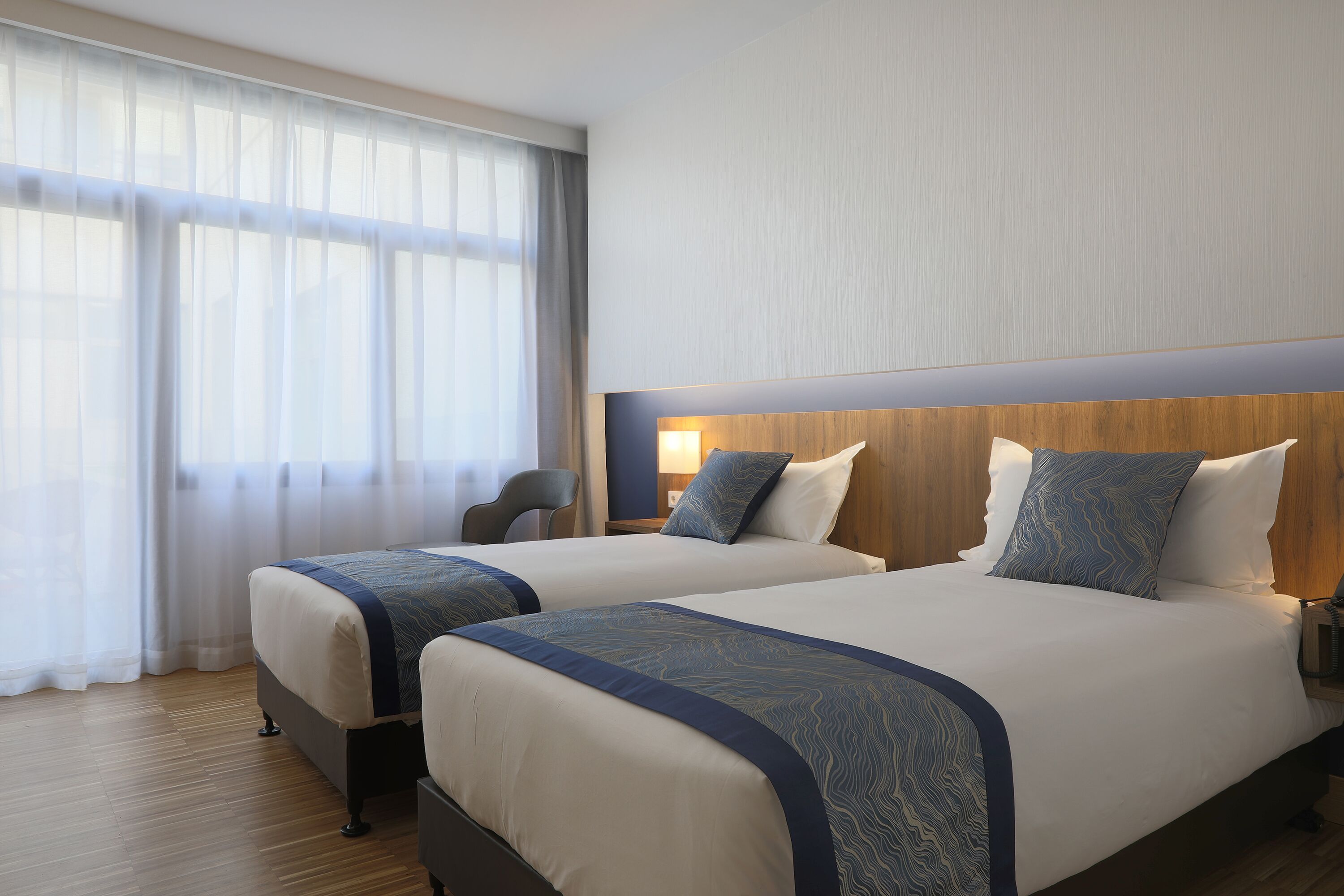 A hotel room with two beds, a wooden headboard, a nightstand, a lamp, and a chair.
