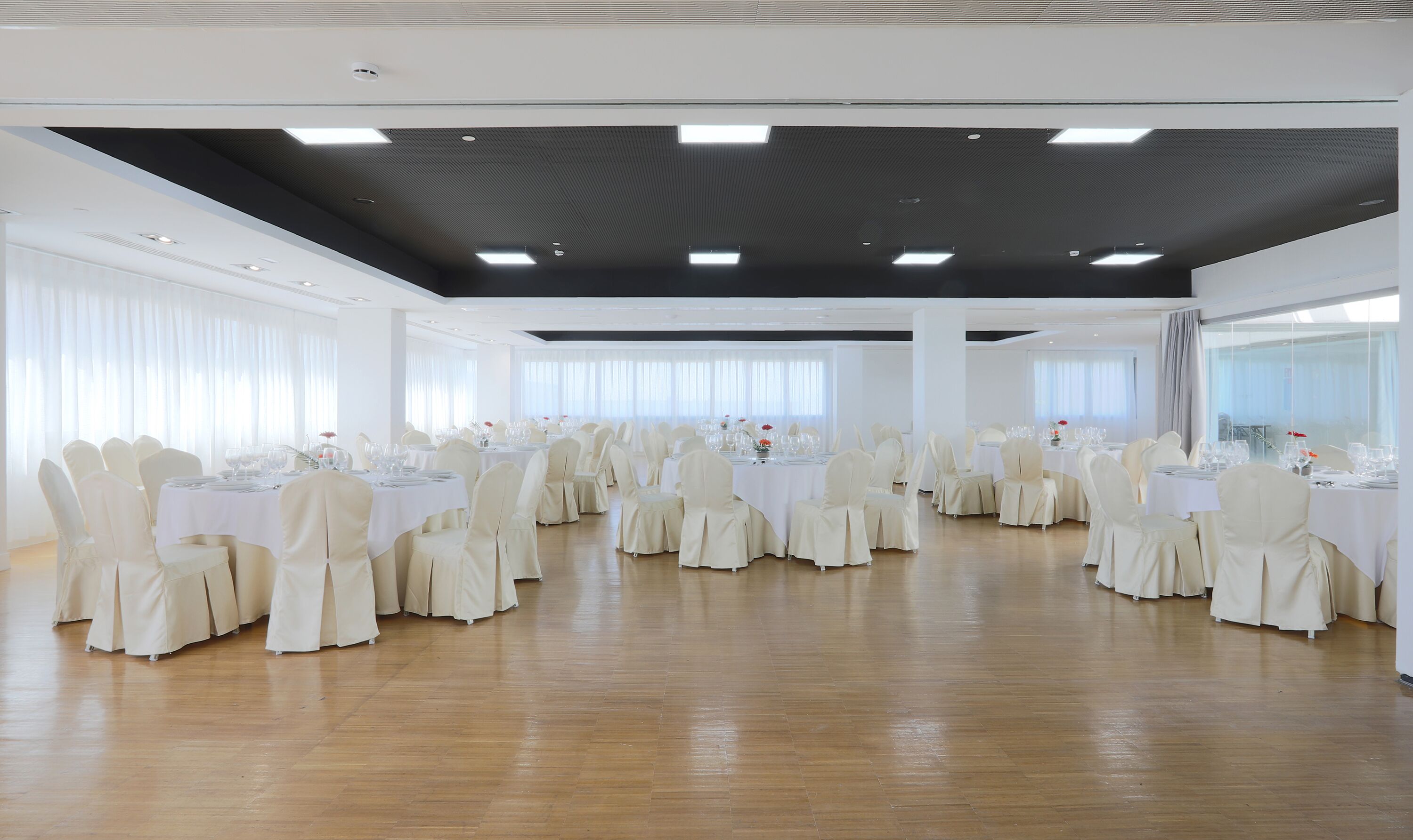 A large, well-lit banquet hall with multiple round tables set for dining, white chairs, and a polished wooden floor.