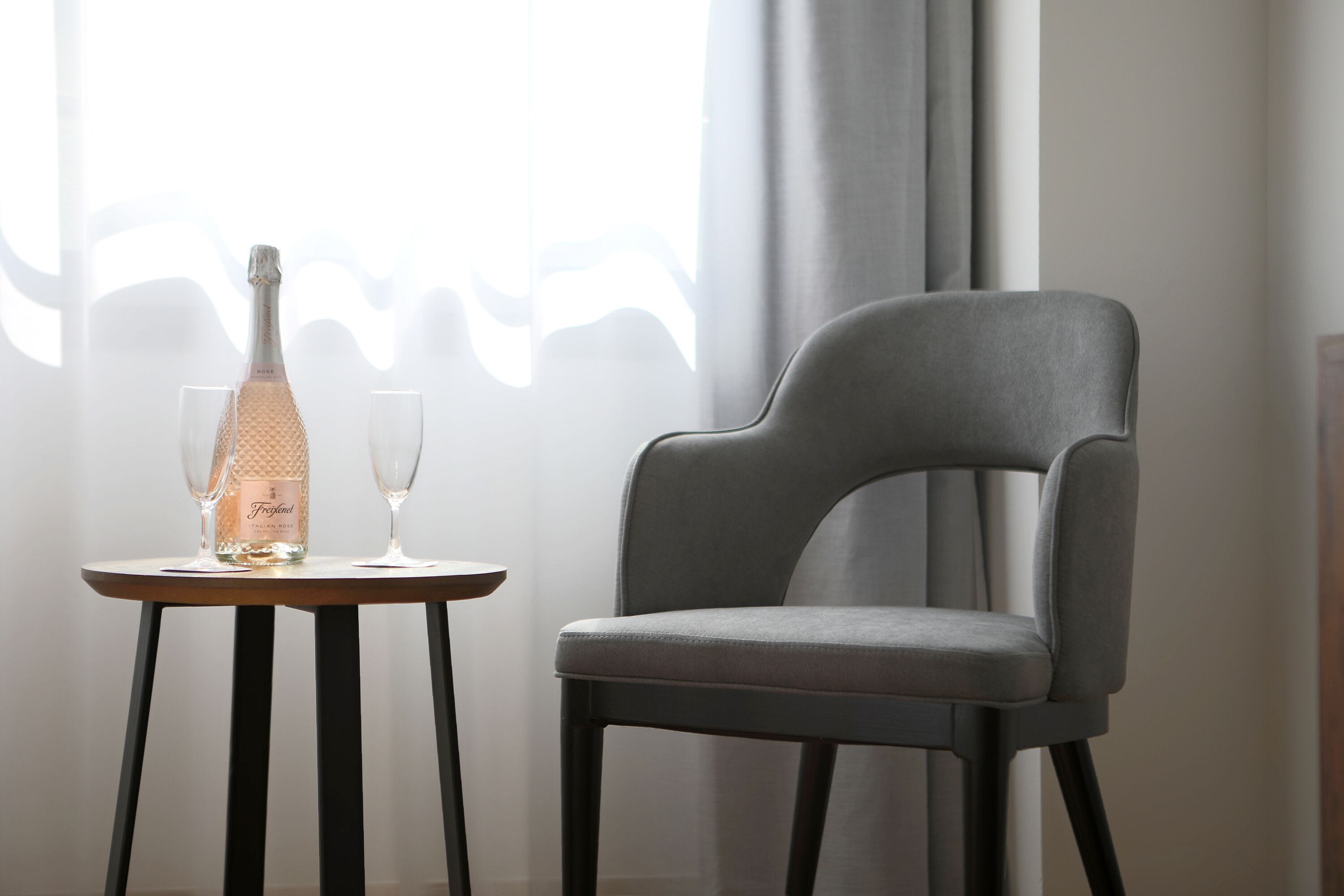 Une pièce moderne avec un fauteuil à revêtement gris et une petite table ronde. Une bouteille de champagne et deux verres sont posés sur la table.