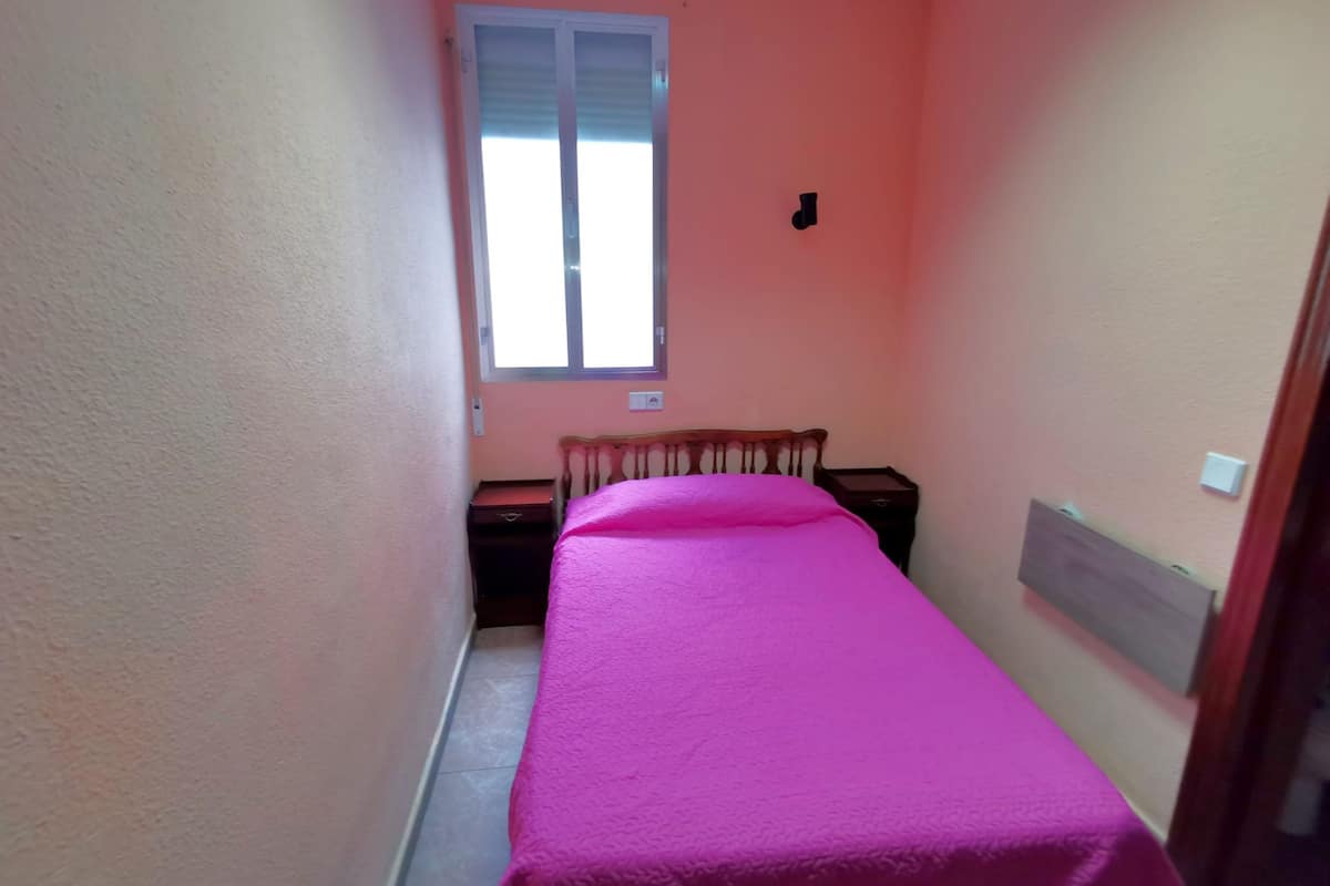 A narrow room with a bed, a nightstand, and a window.