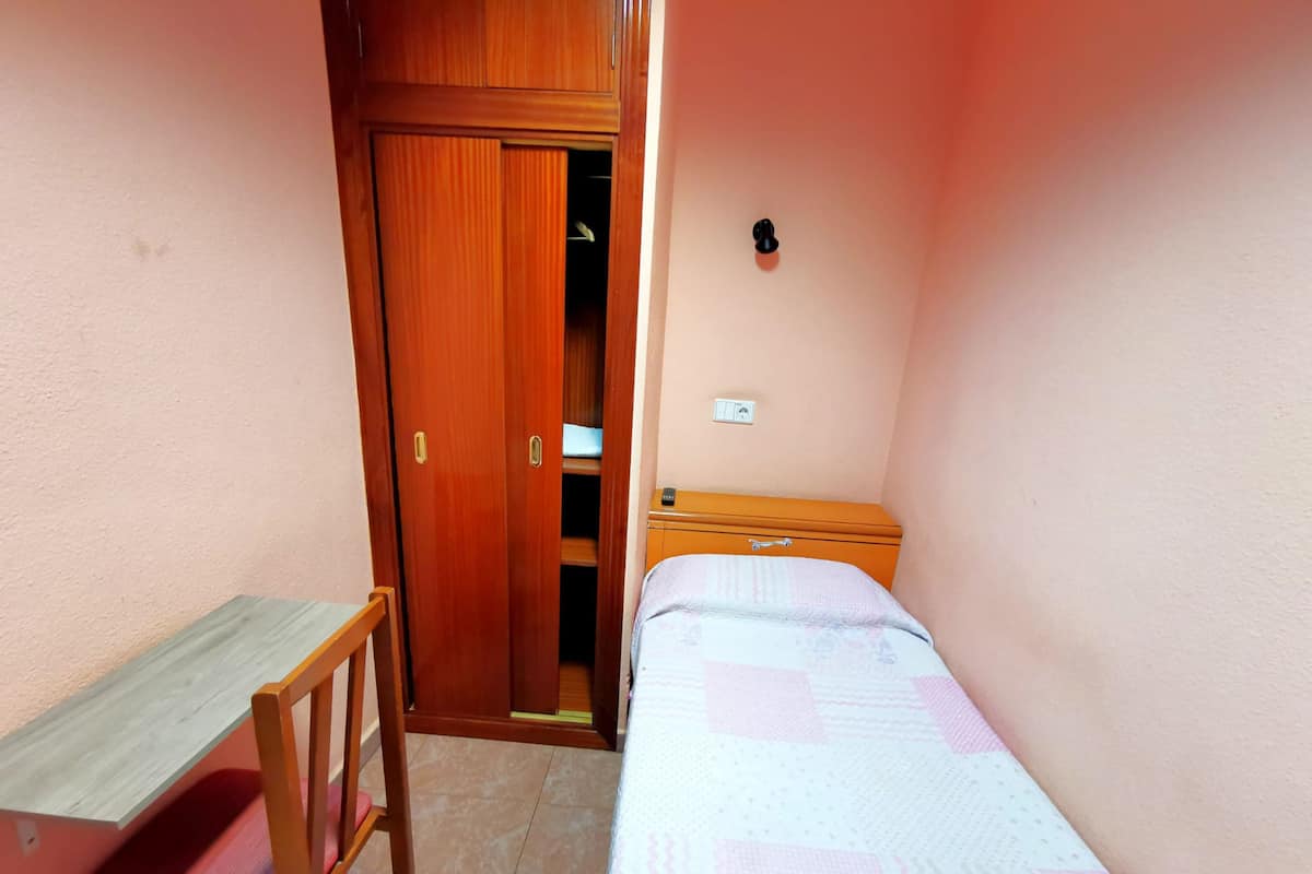 A small bedroom with a wooden wardrobe, a bedside table, a chair, and a bed with a patterned bedspread.