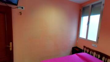A bedroom with peach walls, a bed with a pink blanket, a television on the wall, and a window with blinds.