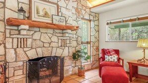 A stone fireplace with a metal screen, a red chair with a patterned cushion, and a window with blinds. - Rosemary Cabin I Couples, Stargazing, Hill Country (Wimberley)