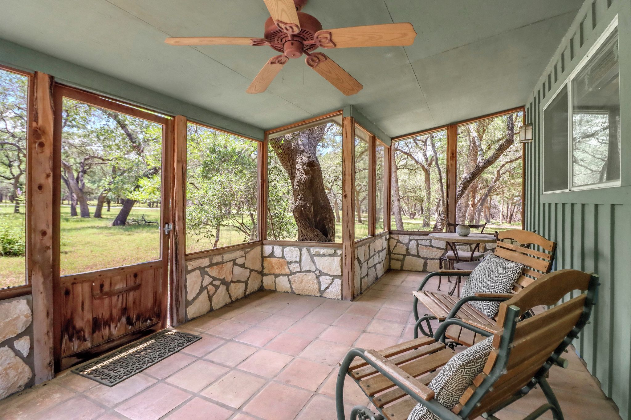 A covered patio with stone walls, wooden furniture, and a ceiling fan.