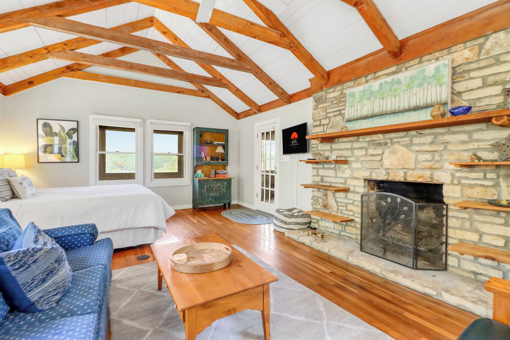 A cozy room with a bed, blue patterned sofa, wooden coffee table, stone fireplace, and wooden beams on the ceiling.