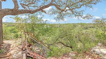 A rustic outdoor area with a metal staircase leading up to a wooden structure, surrounded by dense greenery and a clear sky.
