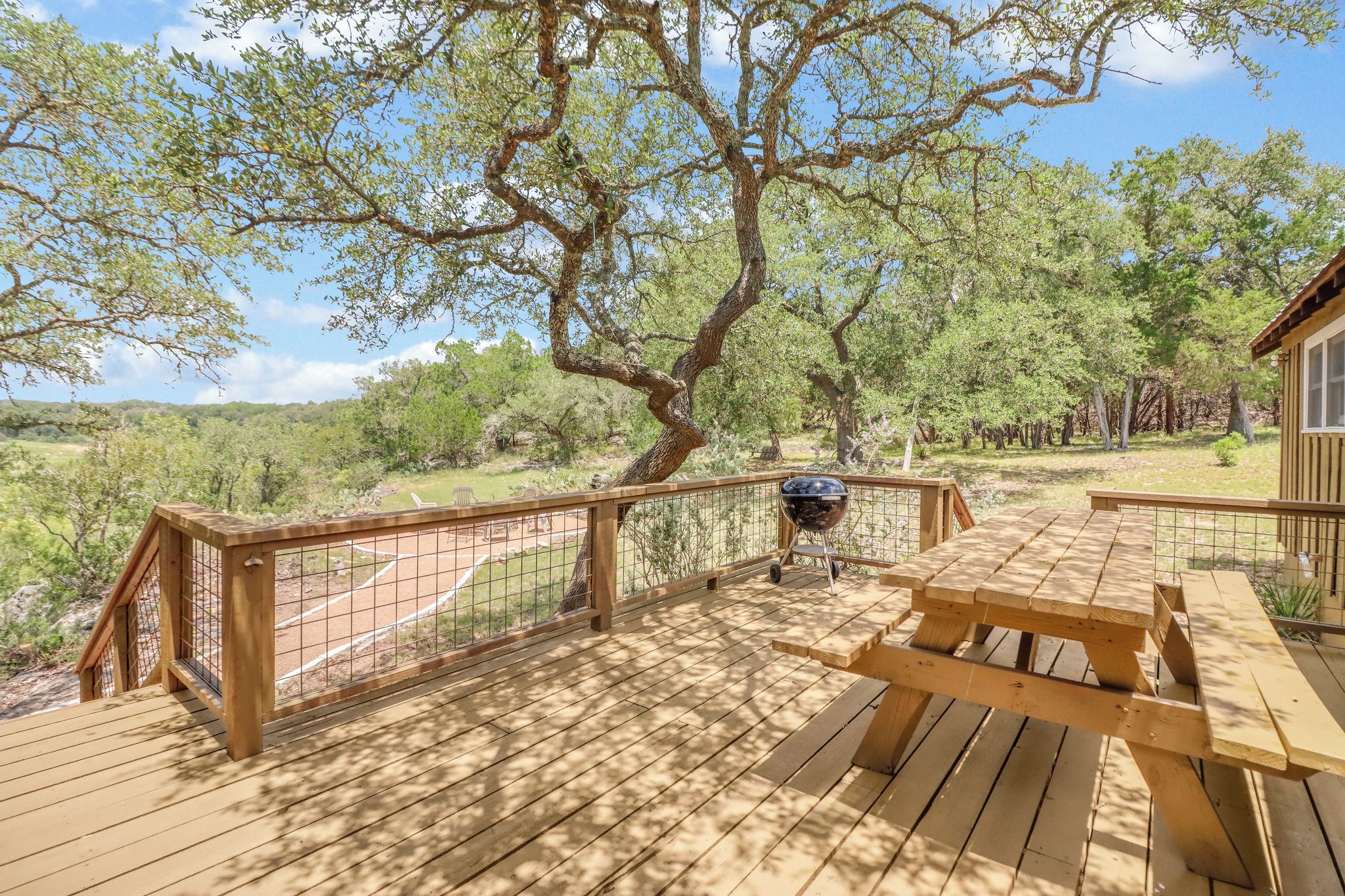 A wooden deck with a picnic table and grill, surrounded by trees and a fenced area.