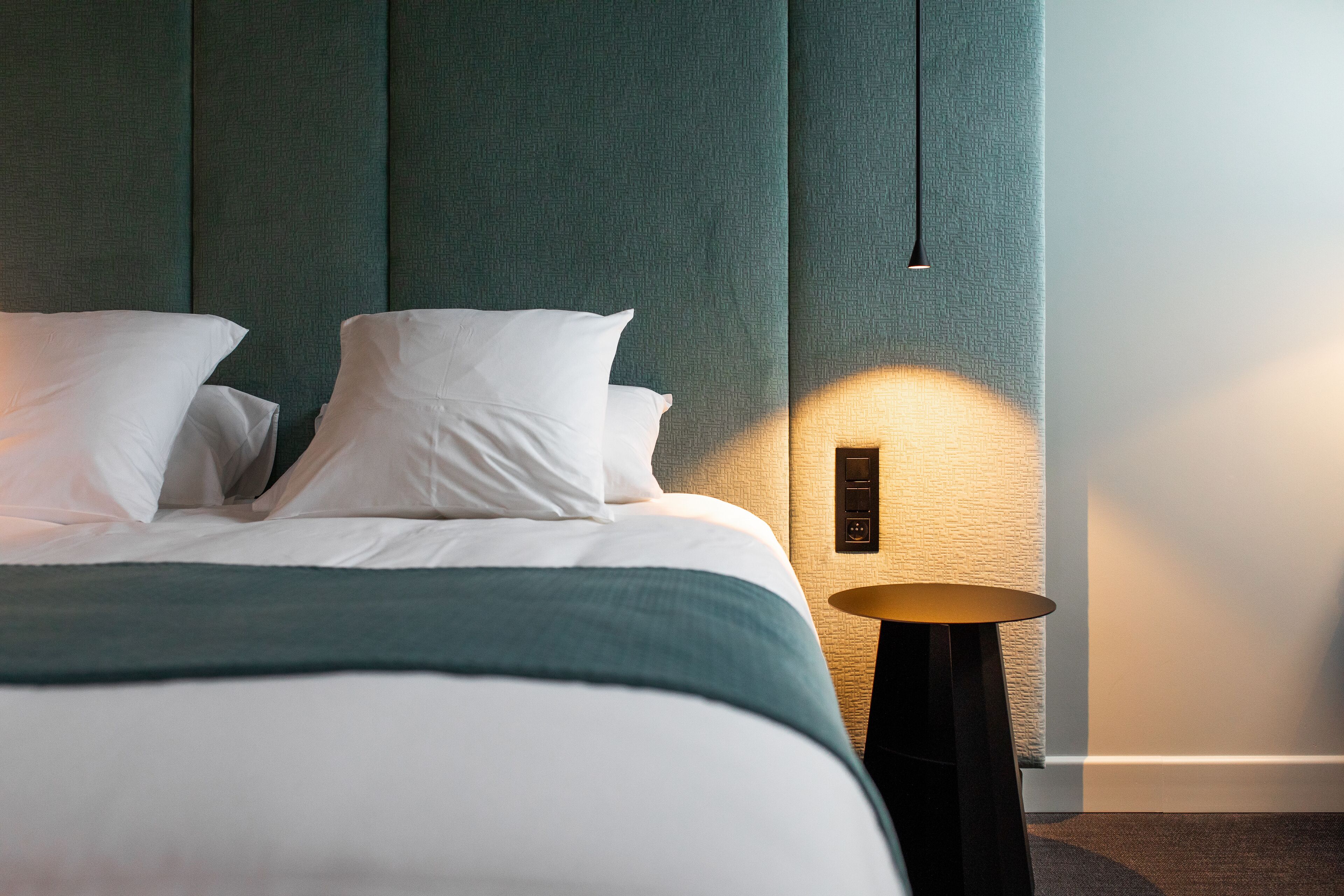A neatly made bed with white linens and a teal throw blanket, a bedside table with a lamp, and a textured wall behind the bed.