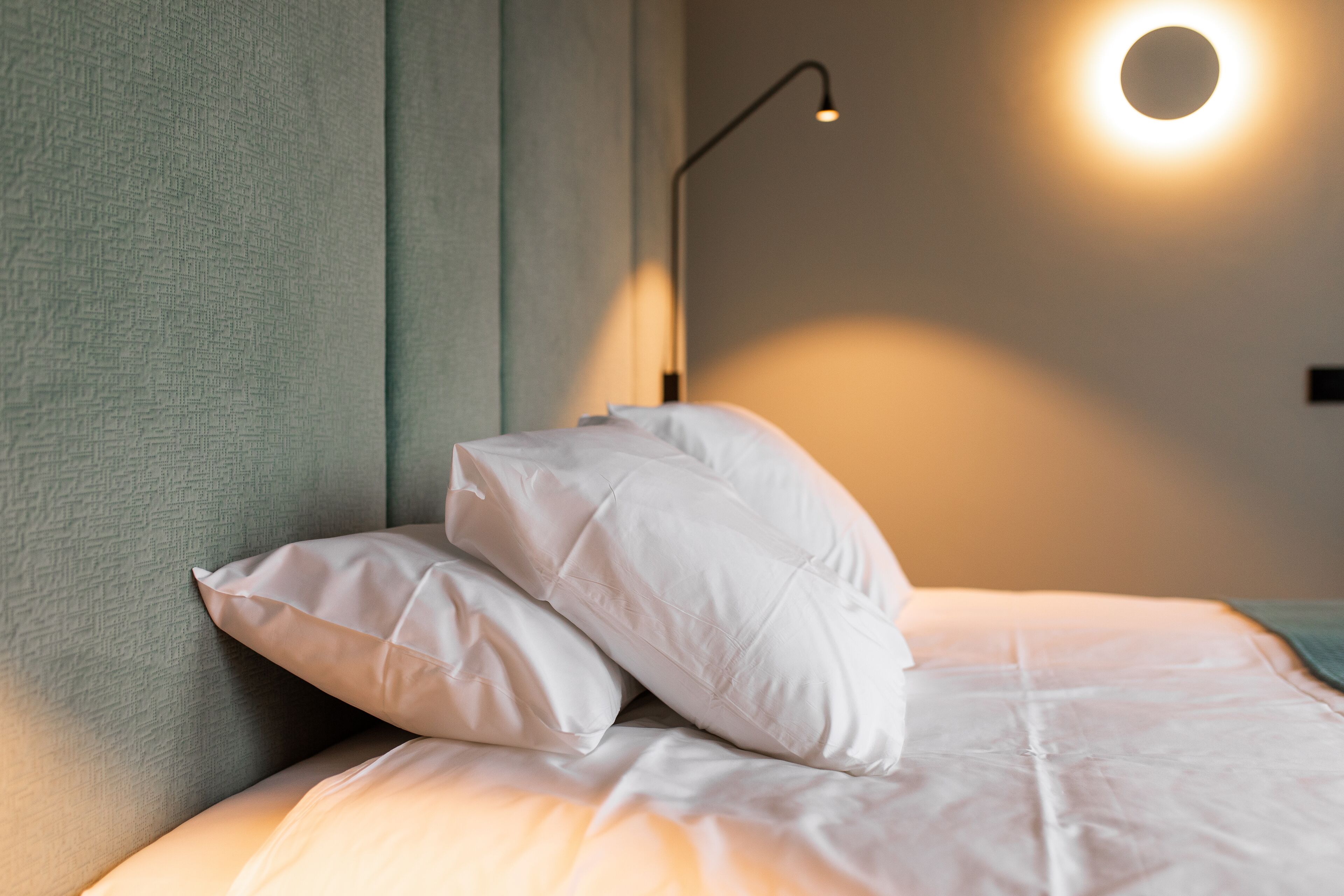 A neatly made bed with white bedding and pillows, a wall-mounted light fixture, and a textured wall.