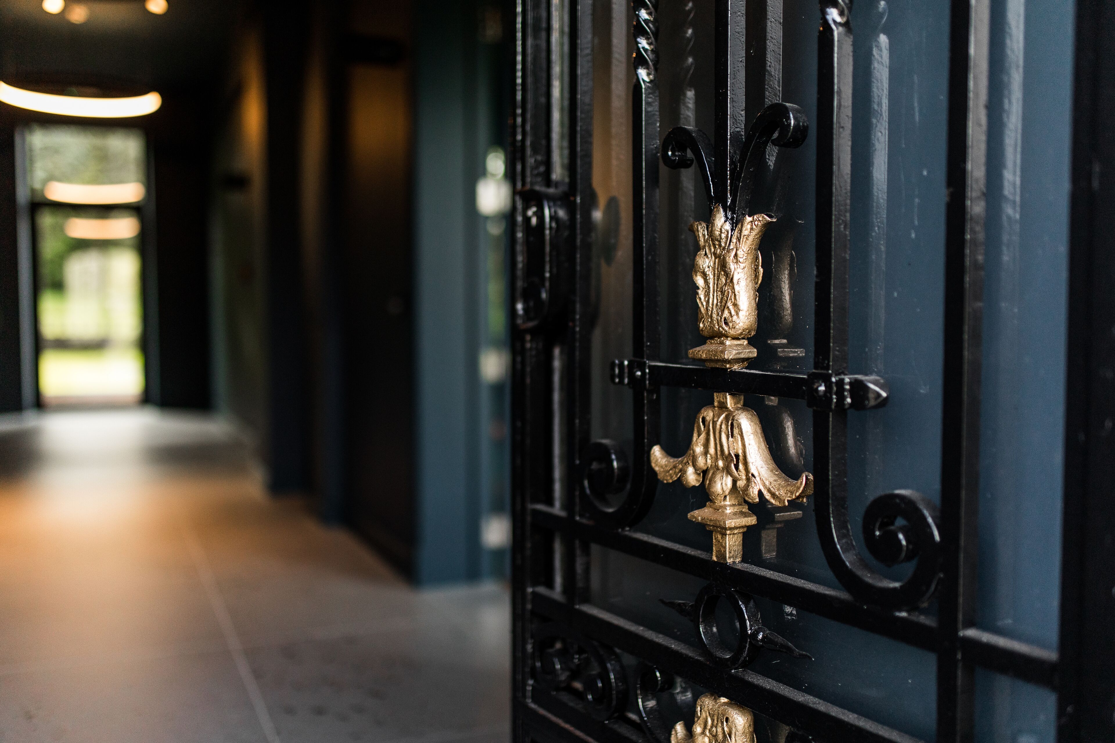 A black wrought iron gate with a golden decorative element, leading to a hallway with a light source at the end.