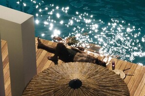 A person lying on a wooden deck by a pool with a straw umbrella.