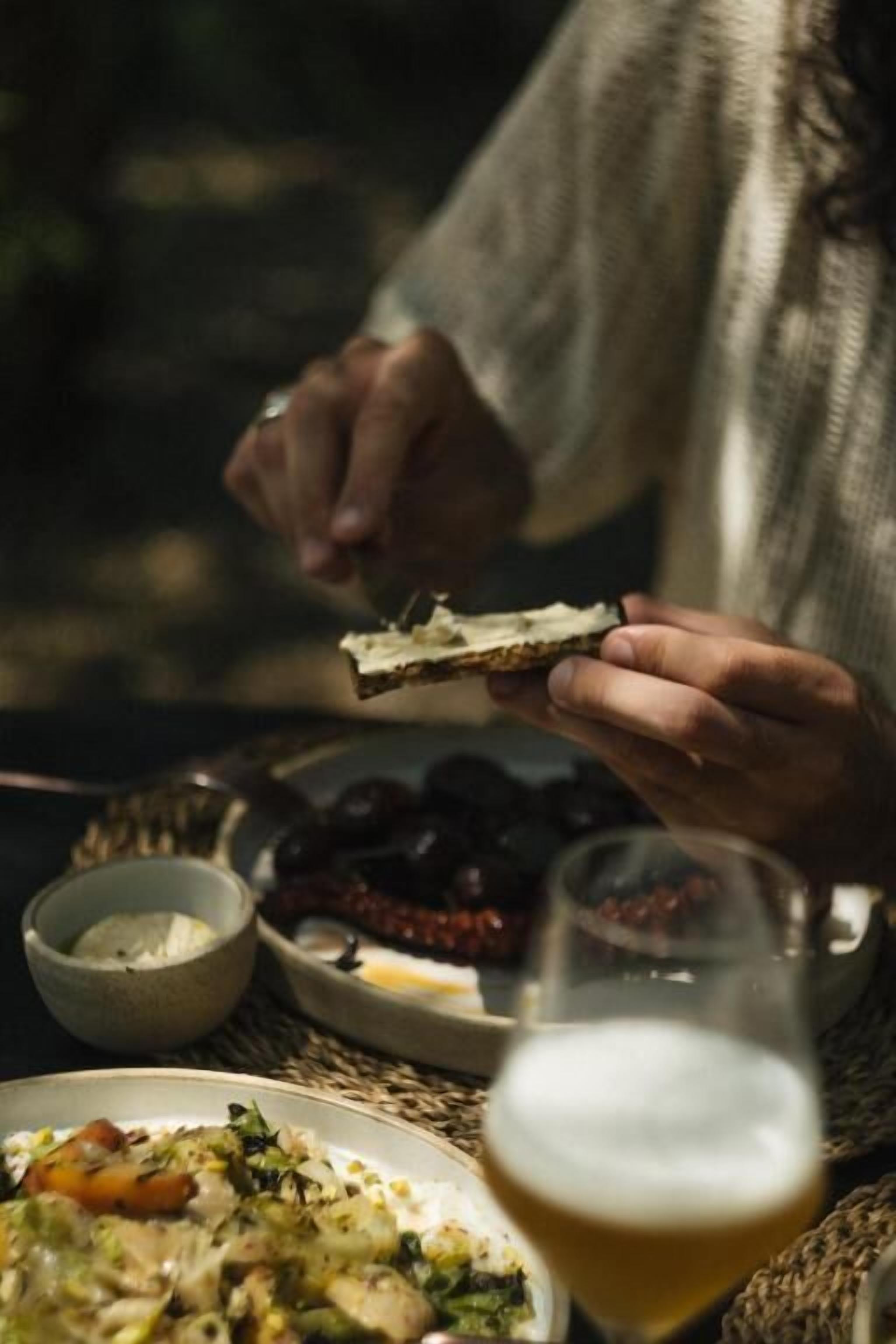 Una persona sosteniendo un trozo de pan, con un plato de aceitunas y una jarra de cerveza sobre la mesa.