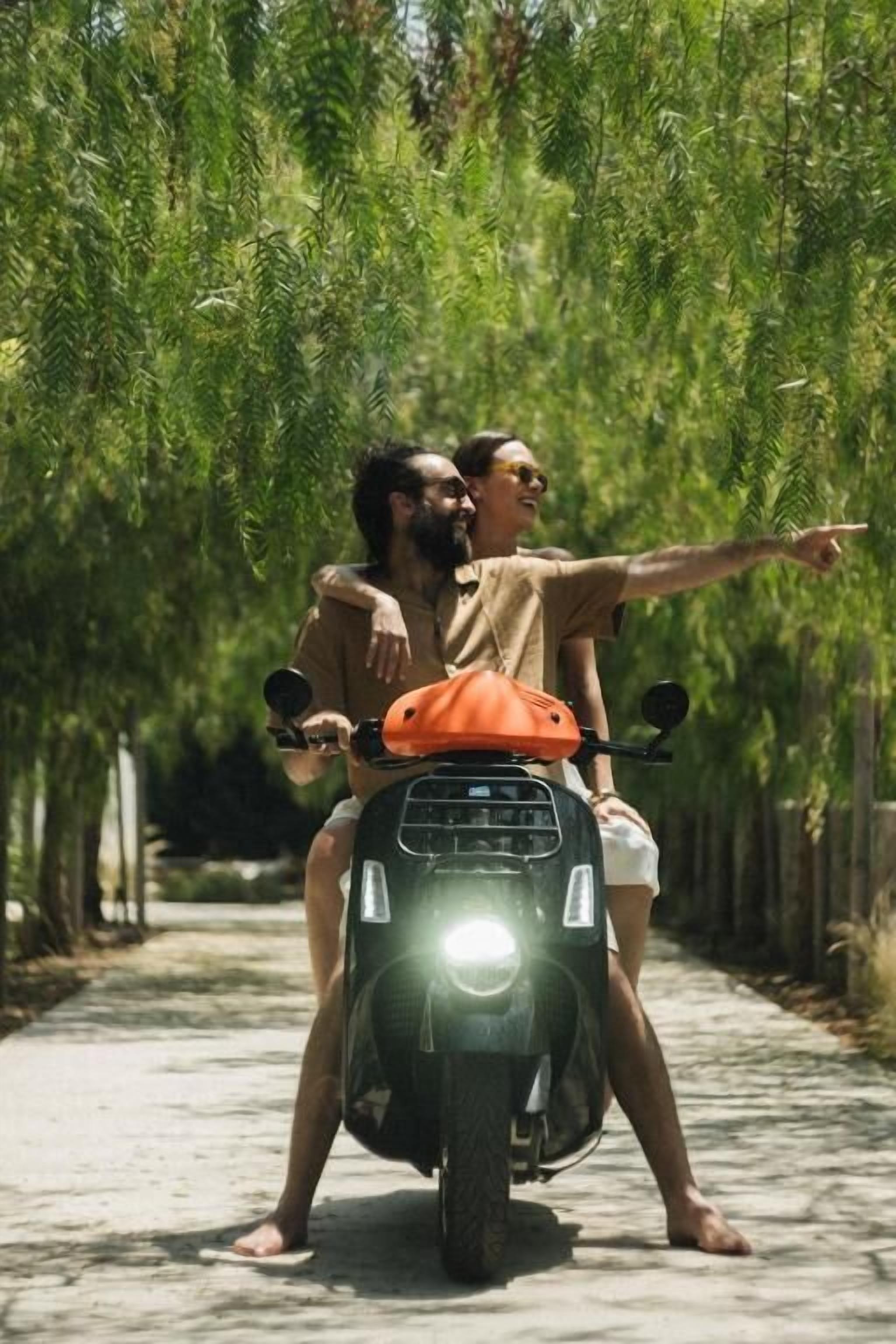Un couple sur un scooter, dans une rue bordée d’arbres.