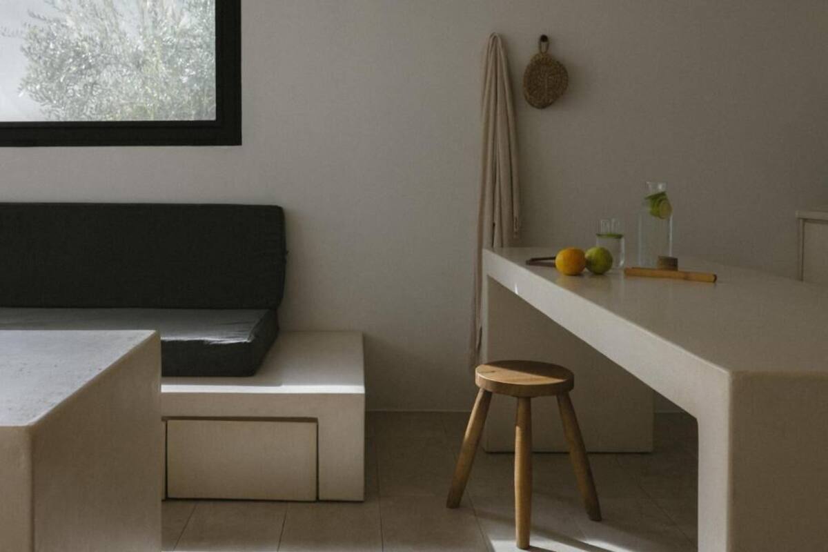 A minimalist room with a sofa, a small stool, a table with fruits and a glass, and a window with a view of trees.