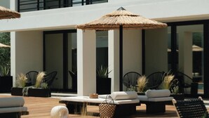 A modern outdoor seating area with lounge chairs and a thatched umbrella, surrounded by potted plants and a wooden deck. - Hooga (Preveza)