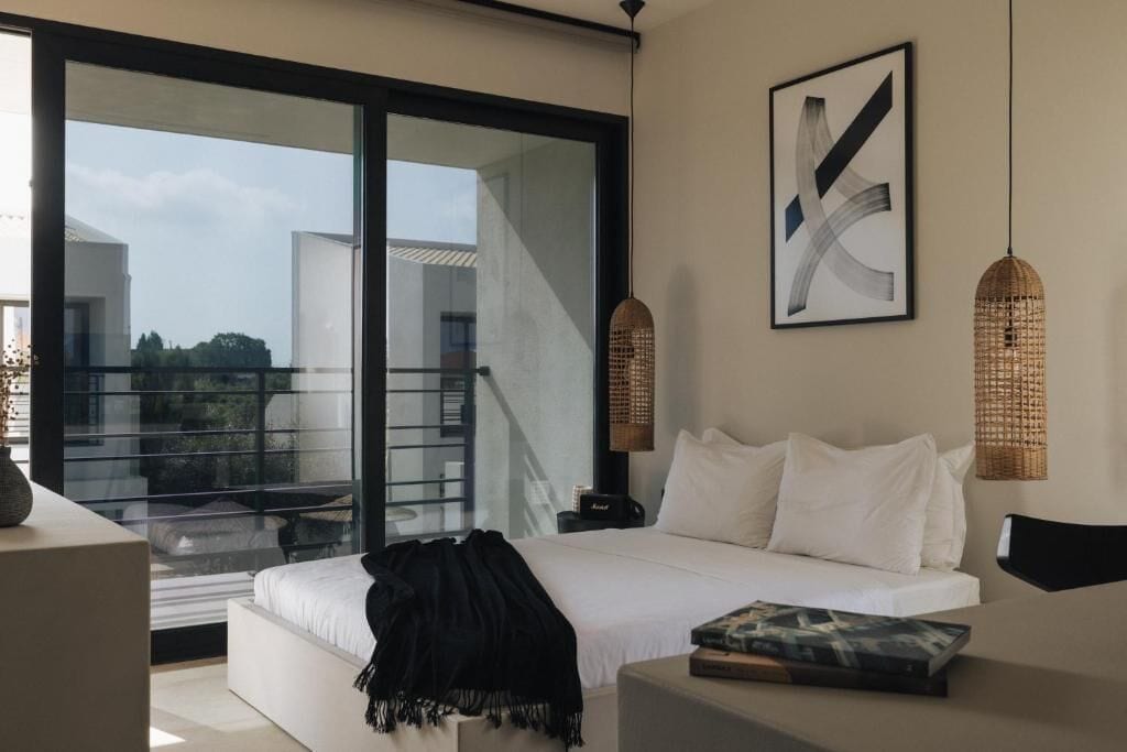 A modern bedroom with a large bed, a glass door leading to a balcony, and a framed abstract painting.