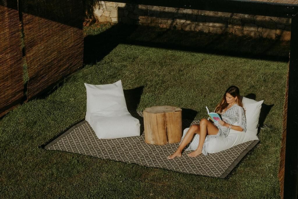 A person sitting on a grass-covered area with a white cushion and a wooden stool, reading a book.