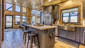 A wooden cabin interior with a kitchen area featuring a large island, bar stools, and a view of the outdoors through multiple windows.