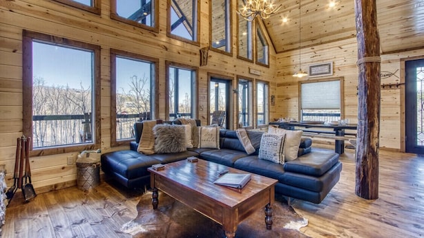 A log cabin interior with a large sectional sofa, a wooden coffee table, and multiple large windows offering views of the outdoors.
