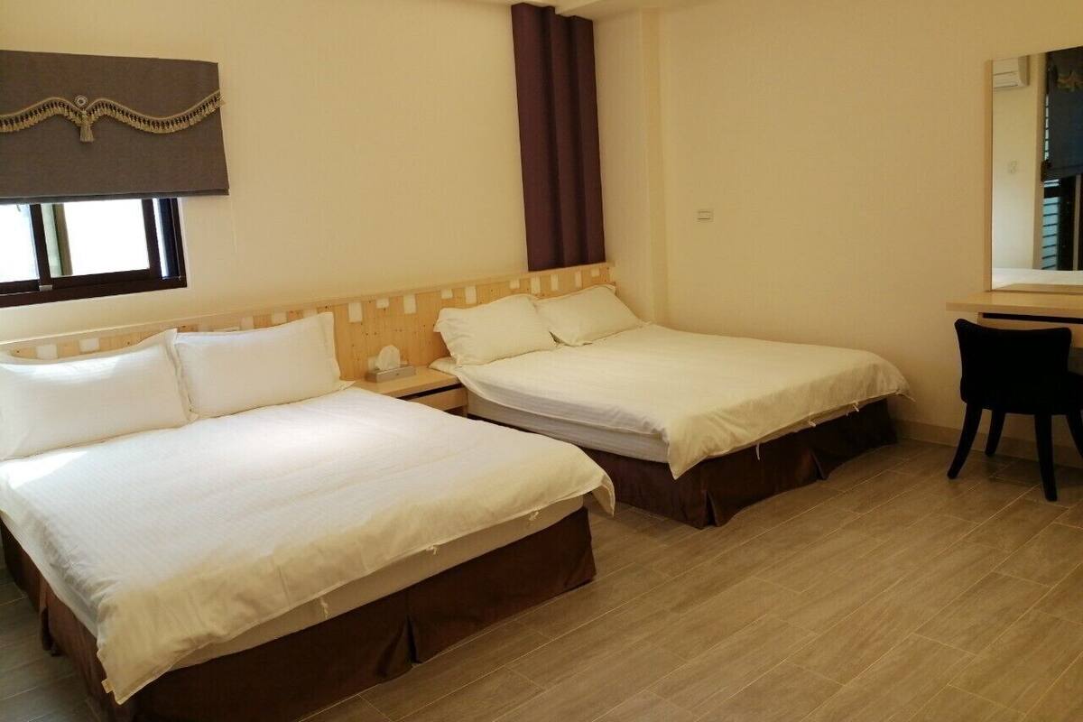 Quarto de hotel com duas camas de solteiro, cabeceira de madeira, escrivaninha com cadeira e janela com cortinas.