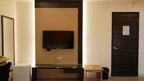 Una sala de estar moderna con un televisor de pantalla plana montado en la pared, una mesita de madera pequeña, un mini refrigerador blanco y una puerta de madera oscura.