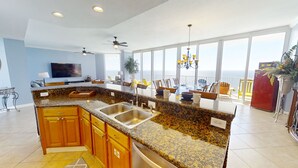 Eine moderne Küche mit Granit Arbeitsplatten, Edelstahlgeräten und Holzschränken. Sie verfügt über eine große Kochinsel mit Spüle, einen Essbereich mit Meerblick und einen Balkon mit einem roten Kühlschrank.