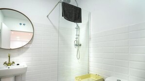 Une salle de bain aux murs recouverts de carreaux blancs, avec un miroir rond, un lavabo doté d’un robinet doré, une douche avec une porte en verre et des toilettes équipées d’un abattant de couleur beige.