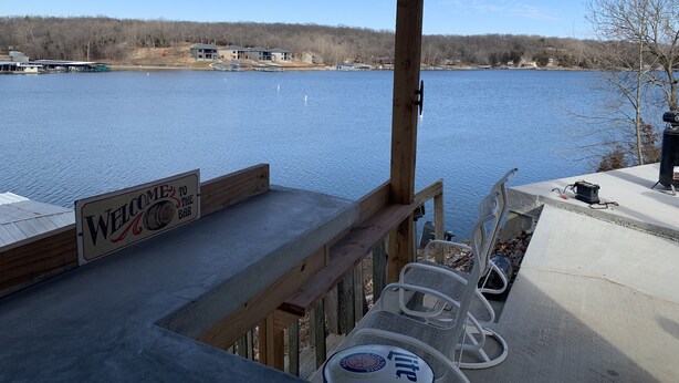 A wooden deck with a 'Welcome to the Bar' sign, outdoor chairs, and a view of a lake with buildings in the background.