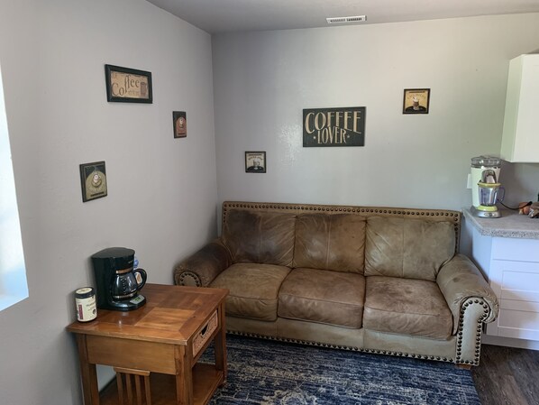 Una sala de estar acogedora con un sofá de cuero marrón, una mesa de centro de madera y una cafetera. Hay cuadros enmarcados en las paredes y una zona de cocina con una licuadora.