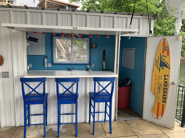 Una pequeña zona de bar con mostrador blanco, dos taburetes azules, una ventana y una decoración con una tabla de surf.