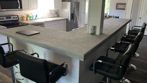 A modern kitchen with white cabinets, a stainless steel refrigerator, a microwave, and a gas stove. There is a bar area with black bar stools and a laptop on the counter.