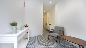 A hotel room with a white desk, a chair, a small table, a potted plant, a fire extinguisher, and a patterned floor.