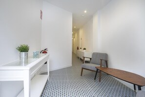 Habitación de hotel con un escritorio blanco, una silla, una mesita, una planta en maceta, un extintor y un suelo estampado.