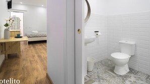 Une chambre avec un plancher en bois, un lit, une table de chevet ornée d’un vase et une fenêtre donnant sur un beau paysage. La salle de bain est équipée de toilettes, d’un lavabo et de carreaux décorés.