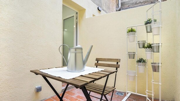 Un petit patio extérieur avec un arrosoir en métal, un banc en bois et un présentoir à plantes en métal, sur lequel sont disposées des plantes en pot.