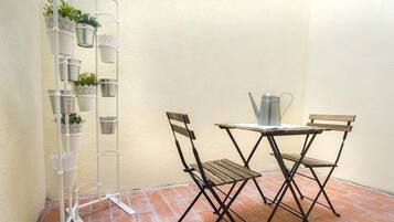 A small patio area with a metal table and two wooden chairs, a metal watering can, and a white plant stand with multiple pots.