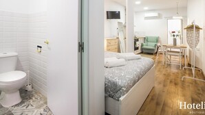 Moderni hotellihuone, jossa on kylpyhuone, kuviollinen päiväpeiteellä varustettu sänky, vihreä nojatuoli, pieni pöytä maljakko täynnä kukkia ja seinälle ripustettu naulakko.