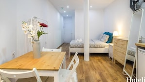 Kompakti hotellihuone, jossa on puinen ruokapöytä, valkoiset tuolit, kuviollinen päiväpeite, yöpöytä lampun kanssa ja suuri peili.
