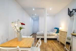 Une chambre d’hôtel compacte comprenant une table à manger en bois, des chaises blanches, un lit avec une couette à motifs, une table de chevet avec une lampe et un grand miroir.