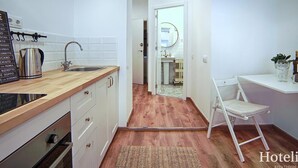 Une cuisine compacte avec un plan de travail en bois, une bouilloire et un évier. Une petite chaise blanche est placée près d’une table blanche, sur laquelle se trouvent une plante et une bouteille.