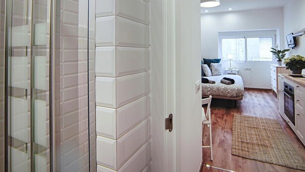 Une chambre d’hôtel moderne dotée d’une douche en verre, d’un lit avec une literie à motifs, d’un petit bureau et offrant une vue sur l’espace de vie adjacent.
