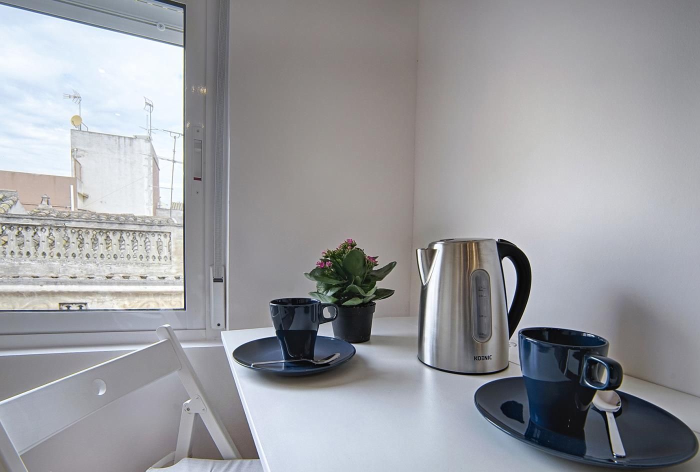 Ett modernt hotellrum med en vit säng, ett fönster med utsikt över byggnader, en liten krukväxt, en vattenkokare i rostfritt stål och två blå koppar med tillhörande fat.